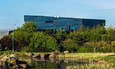 Exterior view of cuboid building set in tree lined gardens, on a sunny day.