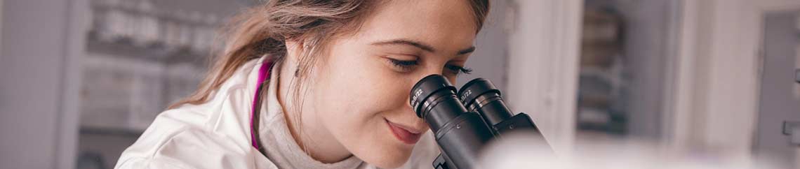 Close up of a female, in a lab coat, looking down a microscope.